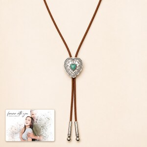 Brown cord bolo necklace with a silver-tone heart slide and turquoise-color center, display view. A personalized greeting-card example appears in the corner.