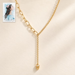 May include: A gold-toned necklace featuring an asymmetrical design with a thick oval link chain on one side and a delicate cable chain on the other. A central vertical drop chain ends in a small gold skull charm with a single embedded clear stone.