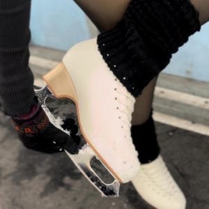 Könnte beinhalten: Weiße Eiskunstlaufschuhe mit einem schwarzen Strick-Beinwärmer. Die Schlittschuhe haben eine goldene Klinge und ein weißes Leder-Obermaterial.
