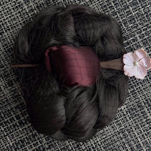 May include: Dark brown hair bun with a burgundy net and a wooden hair stick. The hair stick features a light pink flower with a pearl center. The bun is styled on a textured, dark gray surface.