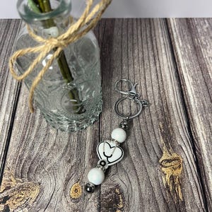May include: White ceramic heart keychain with a black outline and a small black bead. The keychain is attached to a silver metal ring and chain.