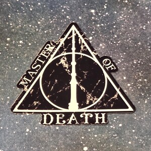 May include: A black and white Deathly Hallows symbol sticker. The sticker features a triangle, a circle, and a vertical line. The words "MASTER OF DEATH" are printed around the triangle. The background is a speckled, dark blue.