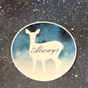 May include: A round sticker featuring a white silhouette of a deer against a blue and white watercolor background. The word "Always" is written in cursive across the deer's body. The sticker is set against a dark blue background with white speckles.