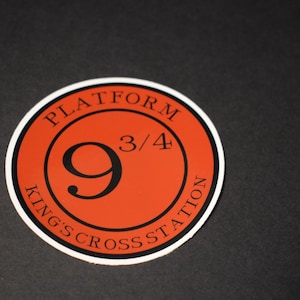 May include: Circular sticker with a white border and an orange center. The sticker features the text "PLATFORM 9 3/4" and "KING'S CROSS STATION" in black, with a large black number "9" in the center. The sticker is on a black surface.