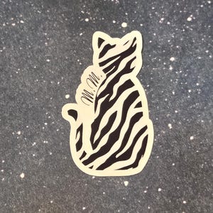 May include: A die-cut sticker featuring a stylized cat silhouette with a zebra stripe pattern in black and white. The sticker includes the initials "M.M." in cursive script on the cat's side.