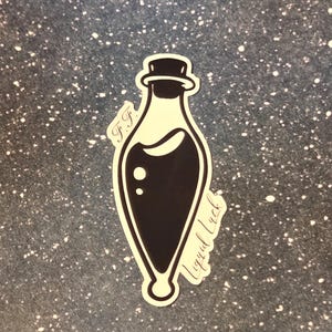 May include: A black and white sticker of a potion bottle with the text "Liquid Luck" and "F.F.". The bottle is filled with a dark liquid and has a rounded bottom and a stopper.