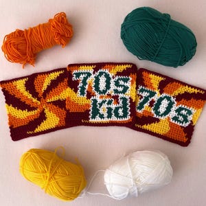 May include: A collection of handmade crochet items with a 70s theme. The image features three square crochet pieces with the words "70s Kid" in green and white. The crochet pieces have a retro color scheme of orange, yellow, and brown. Balls of yarn in orange, yellow, green, and white are also present.