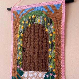 Puede incluir: Tapiz de pared tejido a ganchillo que representa una escena de puerta fantástica. La puerta marrón está enmarcada por un arco marrón con flores amarillas y enredaderas verdes. La pieza está bordeada en rosa y tiene un cielo azul.