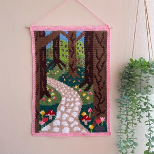 May include: A rectangular crochet wall hanging featuring a forest scene with a winding white stone path, brown tree trunks, green foliage, and red mushrooms. The tapestry is framed by a pink border and suspended from a wooden dowel with pink yarn.