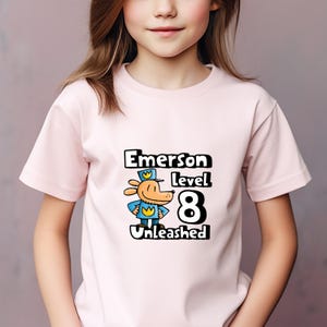 May include: Pink t-shirt with a cartoon dog wearing a blue uniform and a yellow crown. The text "Emerson Level 8 Unleashed" is printed on the shirt.