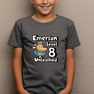 May include: A dark gray t-shirt with a cartoon dog wearing a blue uniform and a yellow crown. The text "Emerson level 8 Unleashed" is printed on the shirt.
