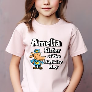 May include: Pink t-shirt with a cartoon dog wearing a blue uniform and a yellow crown. The text on the shirt reads "Amelia Sister of the Birthday Boy".