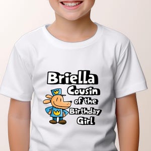 May include: White t-shirt with a cartoon dog wearing a blue uniform and a yellow crown. The text "Briella Cousin of the Birthday Girl" is printed on the shirt.