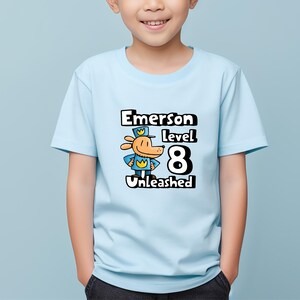 May include: Light blue t-shirt with a cartoon dog wearing a blue uniform and a yellow crown. The text "Emerson level 8 Unleashed" is printed on the shirt.