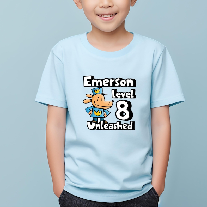 May include: Light blue t-shirt with a cartoon dog wearing a blue uniform and a yellow crown. The text "Emerson level 8 Unleashed" is printed on the shirt.
