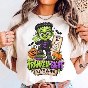 May include: A cream-coloured t-shirt featuring a cartoon Frankenstein monster wearing glasses, standing on books next to pumpkins and an eye chart. The graphic includes the text "FRANKEN-SIGHT" and "EYE'M ALIVE" with small bats and stars.