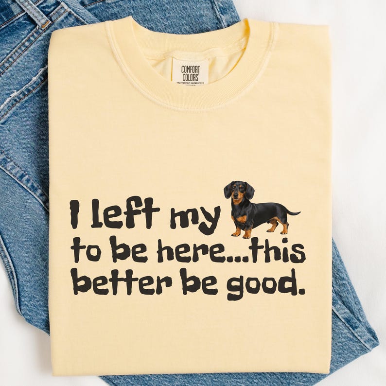May include: A light yellow Comfort Colors t-shirt with the text "I left my to be here...this better be good." and a dachshund graphic. The shirt is folded on top of blue denim jeans.