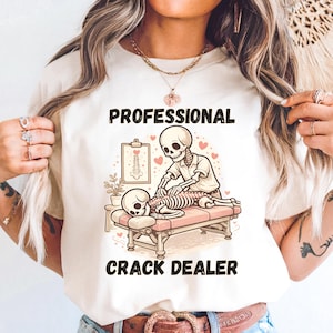 May include: A cream-colored t-shirt featuring a cartoon illustration of skeletons. One skeleton is giving another a back adjustment on a massage table. The text "PROFESSIONAL CRACK DEALER" is printed above and below the illustration.