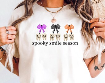 May include: A cream-coloured t-shirt with a Halloween design. The graphic shows three sets of teeth with spooky faces, each with a bow in purple, black, and orange. The text "spooky smile season" is printed below the teeth.