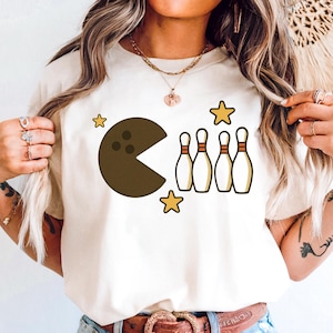 May include: Cream-colored t-shirt featuring a Pac-Man-inspired bowling ball design with a bite taken out of it, alongside four bowling pins and three yellow stars. The graphic is in shades of brown, yellow, and white.