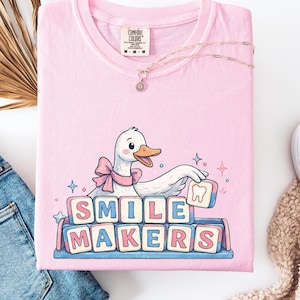 May include: A pale pink t-shirt featuring a cartoon goose with a pink bow, holding a block with a tooth. The text "SMILE MAKERS" is displayed in block letters. A gold necklace is draped across the shirt. The t-shirt has a Comfort Colors label.