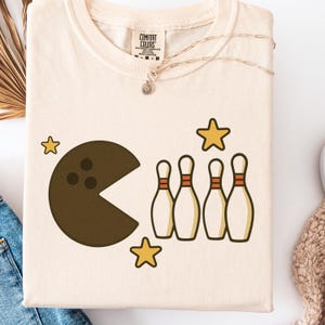 May include: Cream-colored t-shirt featuring a Pac-Man-inspired bowling ball design with bowling pins and stars. The shirt has a Comfort Colors tag. A gold necklace is draped across the shirt.