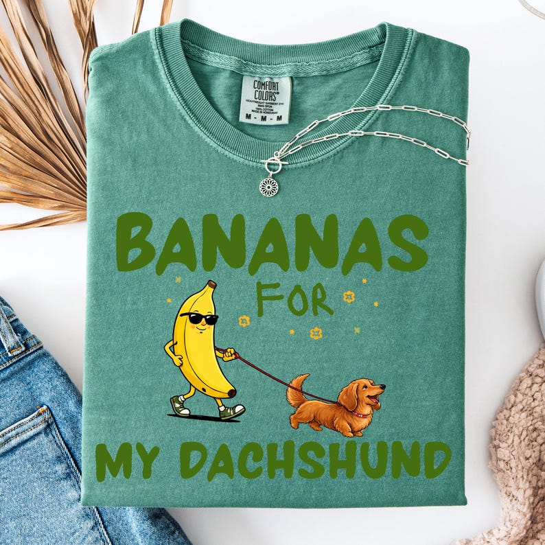 May include: Green t-shirt with the text "BANANAS FOR MY DACHSHUND." The shirt features a cartoon banana wearing sunglasses and walking a brown dachshund on a leash. The shirt has a crew neck and short sleeves.