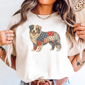 May include: A cream-colored t-shirt featuring a colorful illustration of a dog. The dog's fur is designed with a patchwork pattern of red, white, and blue stars and stripes, resembling the American flag. The dog is smiling with its tongue out.