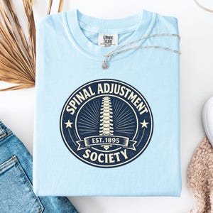 May include: A light blue t-shirt with a circular graphic that reads "SPINAL ADJUSTMENT SOCIETY EST. 1895". The graphic features a spine illustration. A silver necklace with a small pendant rests on the shirt.