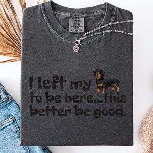 May include: Dark gray Comfort Colors t-shirt with the text "I left my to be here...this better be good." A dachshund dog graphic is on the right. A silver necklace with a pendant is on the shirt.