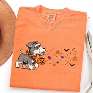 Puede incluir: Una camiseta naranja con el gráfico de un perro schnauzer gris y blanco que lleva un pañuelo con estampado de calabazas y sostiene un cubo de calabaza. El diseño incluye un rastro de puntos con hojas de otoño, murciélagos y una luna creciente.