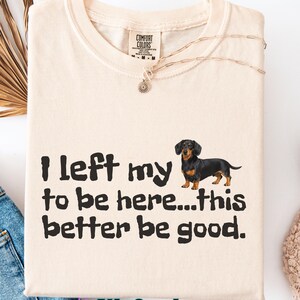 May include: Cream-colored t-shirt with the text "I left my to be here...this better be good." A black and brown dachshund dog illustration is to the right of the text. A gold necklace is draped over the shirt.
