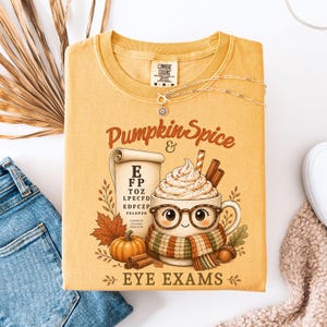 May include: A mustard yellow t-shirt featuring a graphic of a coffee mug with a face, glasses, and a scarf, next to an eye chart, a pumpkin, and cinnamon sticks. The text reads Pumpkin Spice & EYE EXAMS. The shirt has a Comfort Colors brand label.