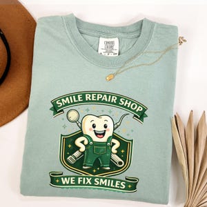 Peut inclure: Un t-shirt vert clair avec un personnage de dent de dessin animé portant une salopette verte et tenant des instruments dentaires. Le t-shirt affiche le texte "SMILE REPAIR SHOP" et "WE FIX SMILES". Un collier en or est drapé sur le t-shirt.