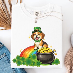 May include: White t-shirt with a cartoon beagle wearing a green leprechaun hat and bow tie, sitting on a rainbow. A pot of gold and shamrocks are also in the design. The shirt has the Comfort Colors brand tag.