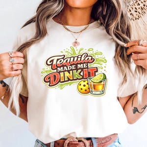 Puede incluir: Camiseta color crema con la frase "Tequila Made Me Dink It" en un gráfico colorido. El diseño incluye un chupito de tequila con una rodaja de lima y una pelota de pickleball. Es una prenda informal.