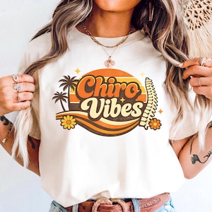 May include: Cream-colored t-shirt featuring a retro design with the words "Chiro Vibes" in orange and yellow. The design includes palm trees, flowers, and a sunburst. The shirt has a crew neck and short sleeves.