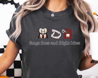 Op de afbeelding: Donkergrijs t-shirt met een Halloween-thema. Het ontwerp bevat een cartoon vampier, tanden en een doosje met flosdraad. De tekst "fangs floss and fright bites" staat eronder.