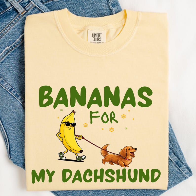 May include: A light yellow t-shirt with the text "BANANAS FOR MY DACHSHUND" in green. The graphic shows a walking banana with sunglasses and a dachshund on a leash. The shirt is folded on a pair of blue jeans.
