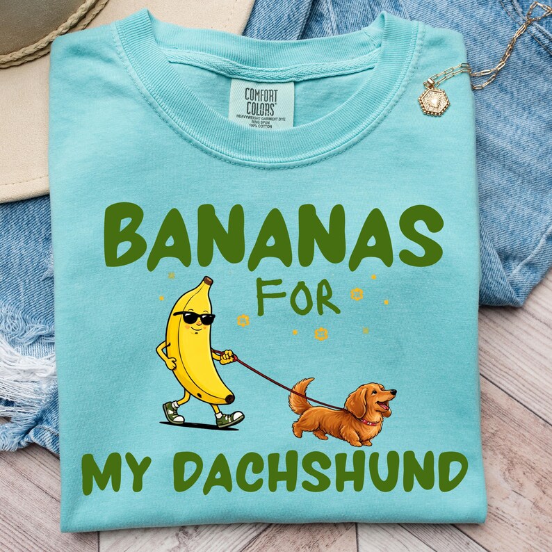 May include: A light blue t-shirt with the words "BANANAS FOR MY DACHSHUND" in green. The graphic shows a banana wearing sunglasses walking a dachshund on a leash. The shirt is made of a soft material.