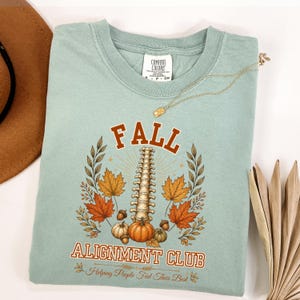 May include: A sage green t-shirt featuring a graphic print with the text FALL ALIGNMENT CLUB and Helping People Feel Their Best. The design includes a human spine, autumn leaves, pumpkins, and acorns in shades of orange, brown, and yellow.