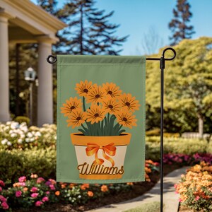 Puede incluir: Una bandera de jardín con un diseño floral. La bandera tiene un fondo verde claro con una maceta de flores naranjas. La maceta es blanca con un lazo naranja y el nombre "Williams" en cursiva.