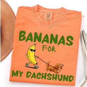 May include: Orange t-shirt with the text "BANANAS FOR MY DACHSHUND" in green. The graphic shows a banana wearing sunglasses walking a dachshund on a leash. The shirt has a Comfort Colors tag.