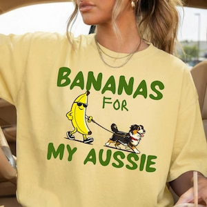 May include: Pale yellow t-shirt featuring a cartoon banana in sunglasses walking a dog. The text "BANANAS FOR MY AUSSIE" is printed in green above and below the graphic.