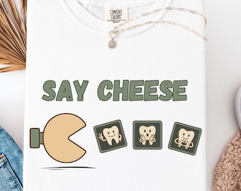 Op de afbeelding: Wit T-shirt met de tekst "SAY CHEESE" in groene letters. Het ontwerp bevat een Pac-Man-achtige vorm en drie vierkante afbeeldingen van tanden met verschillende uitdrukkingen. Een zilveren ketting met een hanger is ook zichtbaar.