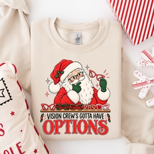 May include: A beige crewneck sweatshirt featuring a graphic of Santa Claus wearing a red hat and green gloves, holding a pair of red glasses. Below him are several pairs of glasses on a shelf with the text "VISION CREW'S GOTTA HAVE OPTIONS".