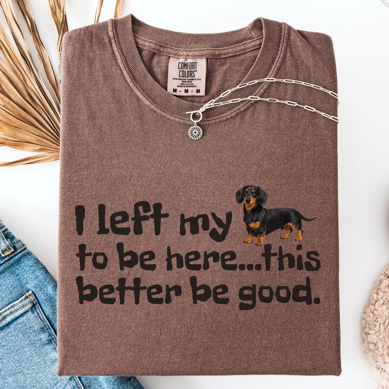 May include: Brown t-shirt with the text "I left my to be here...this better be good." A black and brown dachshund dog graphic is on the right. A silver necklace with a pendant is on the shirt.