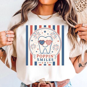 May include: A cream-colored t-shirt featuring a graphic of a tooth wearing heart-shaped sunglasses with American flag designs. The design includes fireworks, stars, and the text "POPPIN' SMILES" in a patriotic theme.