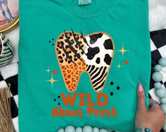 May include: Teal Comfort Colors t-shirt with a large tooth graphic. The tooth design features leopard, cow, and zebra patterns. The text "WILD About Teeth" is printed in orange below the tooth. The shirt is a size XL-TG-FG.