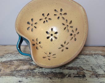 May include: A ceramic berry bowl with a speckled light yellow interior featuring multiple flower-shaped drainage holes. The exterior of the bowl is a deep teal color, and it includes a single curved handle on the side for easy gripping.